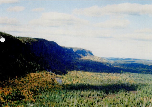 Photograph of the east side of the Kama Cliffs Conservation Reserve. The Jackpine River is running along near the base of the slope. There are cliffs an all sides of this site, with the top forming a relatively flat mesa containing lots of black spruce, white spruce and white birch. 