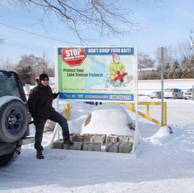 Colour photo of an individual standing in front of a large colourful sign warning anglers not to dump their bait and to protect Lake Simcoe Fishery.