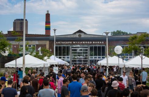 A crowd of people browsing tents set up for a market outside of the Artscape Wychwood Barns building. The building, an old streetcar repair facility, has large windows and a multi-coloured smoke stack.