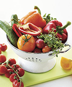 white colander with greenhouse cucumbers, peppers, tomatoes, herbs on a green cutting board