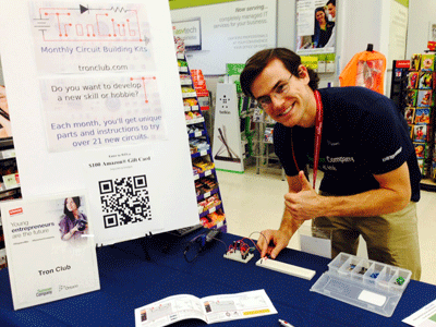 Photo of Greg DeLaunay, founder of Tron-Club, pictured here with an electric circuit board and a sign describing his business.