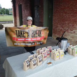 Photo of Kyrstan Edmondson, founder of Take A Break, standing behind her coffee stand holding her business banner.
