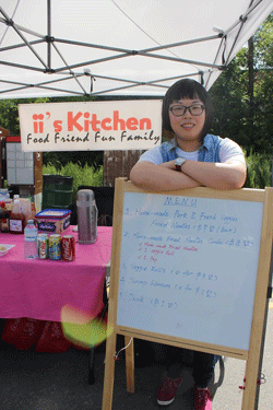 Photo of Yiyi Liang, founder of ii’s Kitchen, leaning on a menu sign in front of her food stand.