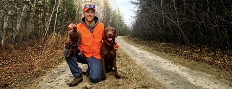Photo of a hunter in an orange vest and his dog. Hunter is holding up a grouse.