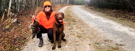 a women dressed in hunter orange holding a grouse with her dog
