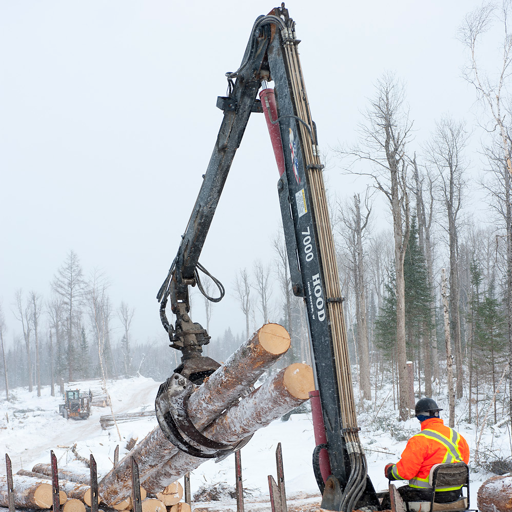 Une personne aux commandes d’une grue de chargement à l’extérieur, en hiver, qui empile des billes dans un camion.