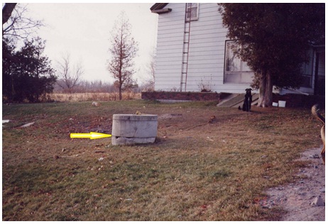 The centre of Figure 11-11 shows the concrete tiles and lid of a dug well. The joint between the first and second concrete tiles is broken and not sealed. The unsealed joint creates a pathway for foreign materials to enter the well. Also a dog, tied to the top of the well cover, is shown on the right side of the photograph near a tree. There is a potential for animal waste to be discharged near or on the well. Animal waste can migrate to and impair the well water used for human consumption.