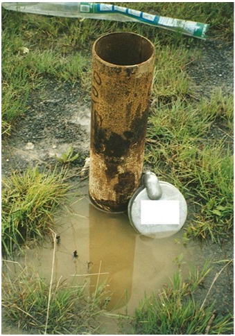 Figure 11-5 shows ponded water that potentially contains pathogens located immediately beside the well. Ponded water can migrate downward through soil pores and bedrock fractures and along the side of the well casing by gravity. The ponded water can enter and impair the quality of the well water. Figure 11-5 also shows an older style non-vermin-proof well cap removed from the well and placed in the ponded water. Any pump equipment installation, repair work or other alterations would require this well cap to be replaced with a vermin-proof well cap.