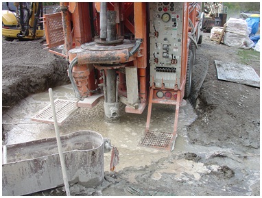 Figure 12-6 shows the back of an orange coloured drilling rig. A black coloured drilled well casing extends out of the muddy water through a rotary table. The drilling rig is holding on to the casing with the rotary table and a welded piece of steel. During drilling the flow around the outside of the casing created a void in the ground. The overburden above the void has collapsed creating a 10 metre (30 foot) deep by 4 metre (13 foot) wide hole filled with water. The void was unknown until the time of overburden collapse. The void created a serious safety issue for anyone working on the back of the drilling rig.