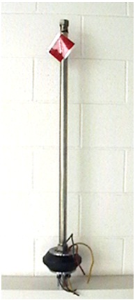 This photograph shows a flowing well packer unit. The unit consists of a packer located at the bottom of a long stainless steel rod. The length of the rod allows the packer to be placed below the frost line. The packer is made out of black coloured Ethylene Propylene Diene Monomer rubber between two stainless steel plates. The bottom of the packer is equipped with an air vacuum valve or a combined air vacuum and air release valve to ensure that air can move into and out of the well but water cannot. The photograph shows the packer area comes equipped with proper gauged electrical cables. Electrical wiring from the submersible pump in the well can be attached to the cables in the bottom of the unit. Electrical cables can be attached to the top of the packer to allow the wiring to extend out of the well and connect to the electrical service. At the top of the steel bar is a nut. A socket wrench can be attached to the nut and can turn the steel bar. When installed in the well, turning the bar squeezes the plates and expands the packer to the sides of the well casing.