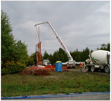 Figure 15-52 shows the cement pump truck with its boom in the air and over the drill rig almost prepared for the pumping of concrete into the flowing well from Figure 15-49. The cement truck is backing up to the cement pump truck to begin the plugging operation.