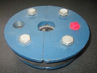 The photograph shows an example of one kind of sanitary well seal. In this case, a blue steel plate is underlain by a black neoprene gasket. The gasket is underlain by another blue steel plate. One waterline can pass through the centre of the well seal. An air vent can be threaded onto the seal on the left side of the well seal. The red plug can be removed on the right side of the well seal to allow for an electrical line or other equipment through the well seal. By tightening the four steel bolts on top of the well seal, the two plates are tightened together expanding the neoprene gasket. The squeezed gasket seals the waterline, electrical line and the casing. The well seal is typically found at the top of the well casing. Other sanitary well seals have two holes for two waterline installations used in jet pumps.