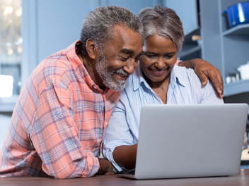 An older adult with their arm around another older adult’s shoulders, looking at a laptop.