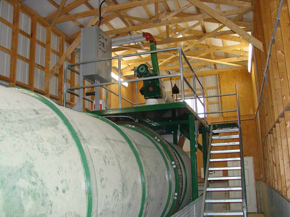 DComposteur à tambour dans un petit local et escaliers à droite menant à l’aire de réception des matières en haut du composteur, où l’équipement est situé.
