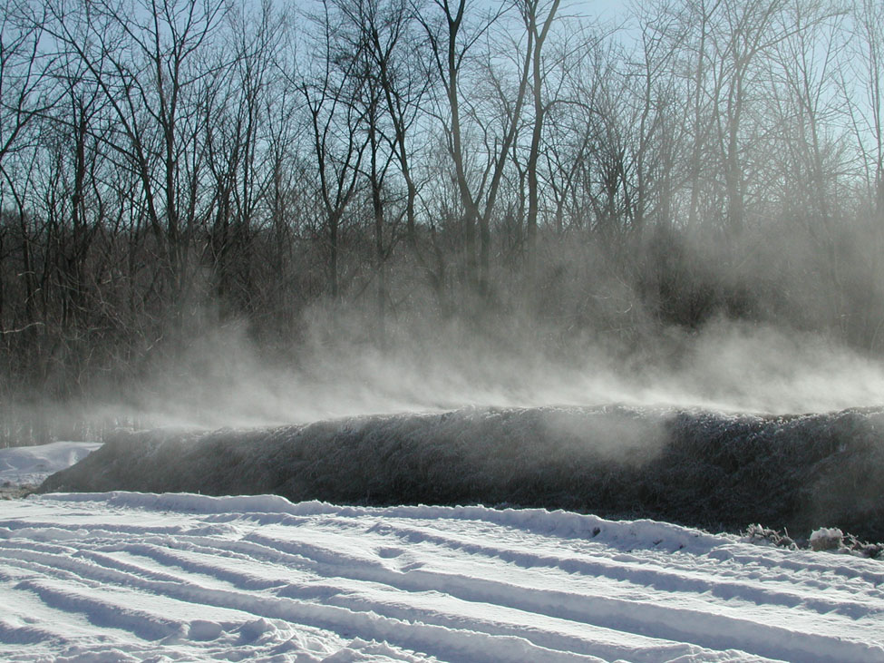 Picture shows a row of composted materials in a filed in the winter. Due to the nature of composting, the windrow is giving off steam in the cold temperature.