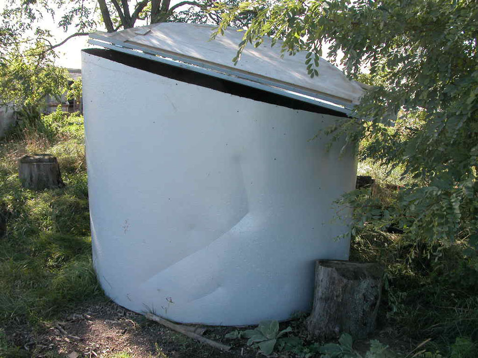 A blue, above ground container with a large lid used for composting of smaller livestock. The container is sitting in a forested area of the farm.