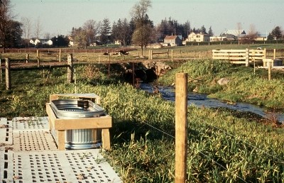 Abreuvoir de remplacement pour le bétail avec socle renforcé, près d'un cours d'eau clôturé. La traverse à mi niveau permet aux animaux d'aller paître des deux côtés du cours d'eau.