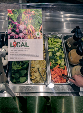 This is a picture of a communication tool placed at St Paul's University College's Wheelock Dining Hall at the University of Waterloo above a salad bar. The communication tool shown is signage about Chartwells Buy Local Program featuring a cornucopia of Ontario vegetables and identifying a local family farm supplying St. Paul's University College. Chartwells is making communication tools co-branded with the Foodland Ontario logo available to colleges, universities and schools to promote local food on their campuses.