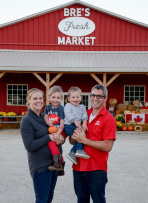 This is a picture of Breann and Kyle Gillespie with their two children standing in front of their store at their on-farm market. Breann and Kyle are the owners of Bre's Fresh Market located near Tillsonburg in Oxford County. 