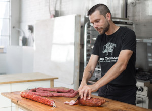 This is a picture of Mike McKenzie with a knife in his hand making hand-crafted artisanal meats. Mike McKenzie is the founder of Seed to Sausage, a company in eastern Ontario that makes artisanal cured meats. 