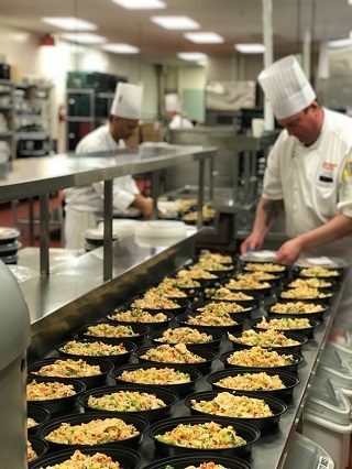 The image shows 3 rows of portioned meals being prepared in a commercial kitchen by a chef. On the left side of the photograph further back from the first chef appears a second chef also preparing portioned meals.