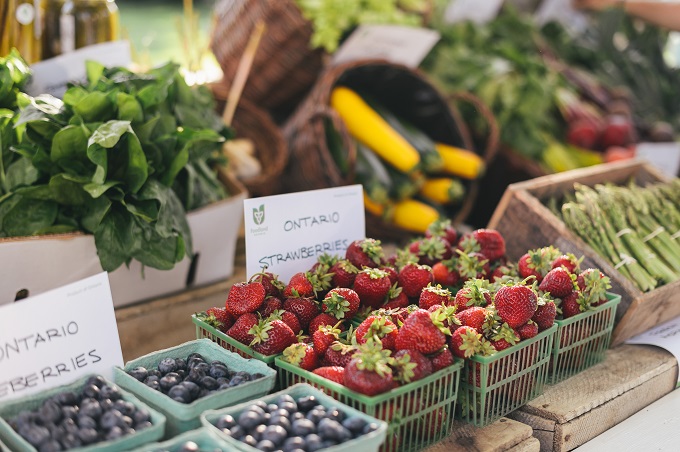 L'image est un marché extérieur avec des bleuets, des fraises et des asperges dans des paniers à l'avant. Des épinards, des courgettes jaunes et vertes sont à l'arrière-plan. Ils sont dans des boîtes et des paniers. D'autres légumes sont sur la table, brouillés derrière eux.