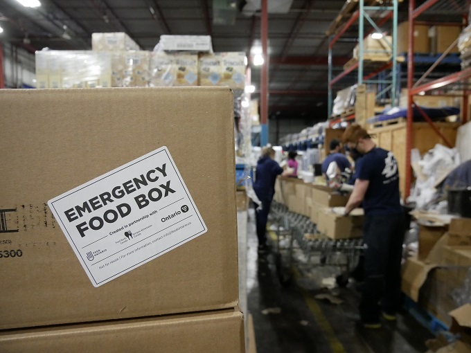 L'image comprend une boîte au premier plan gauche. Un autocollant dit " Emergency Food Box " avec les logos de commanditaires en dessous. À droite de la boîte, en arrière-plan, des bénévoles portant des masques et des gants remplissent des boîtes.