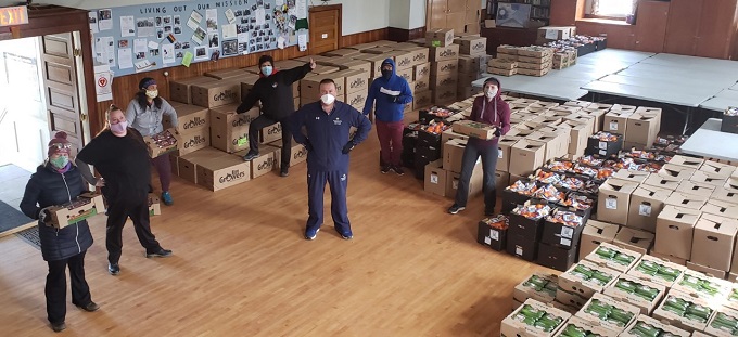 The image shows volunteers wearing masks and gloves - observing social distancing - while packing and carrying boxes of fresh produce for distribution to families in need.