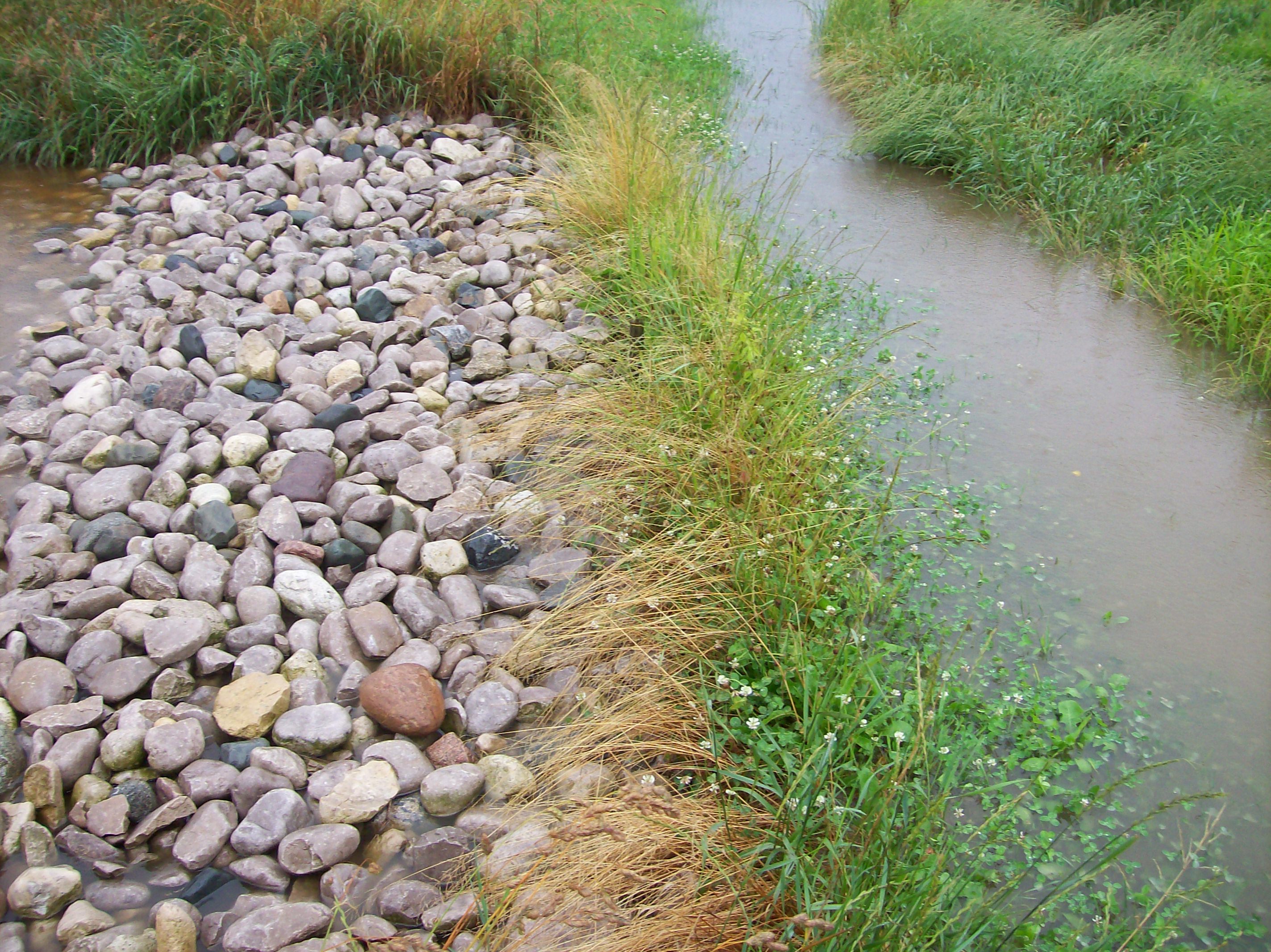 Photo showing the rocked emergency overflow spillway from the holding pond into the vegetated flow path during an extreme runoff event to prevent overtopping of the berm structure. Water in the right side of the photo is stored in the holding pond and is flowing to the left through the rocks to enter vegetated area without causing erosion damage.