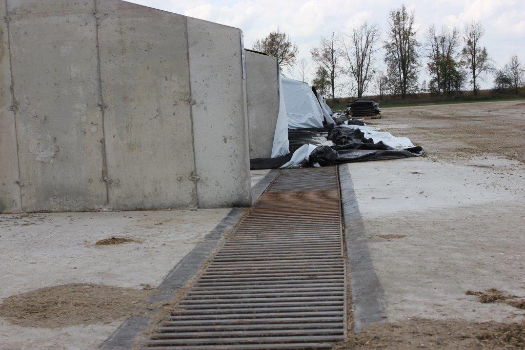 Photo montrant une tranchée de collecte d’effluents recouverte d’une grille métallique perpendiculaire à l’extrémité d’un silo-couloir vide en béton.