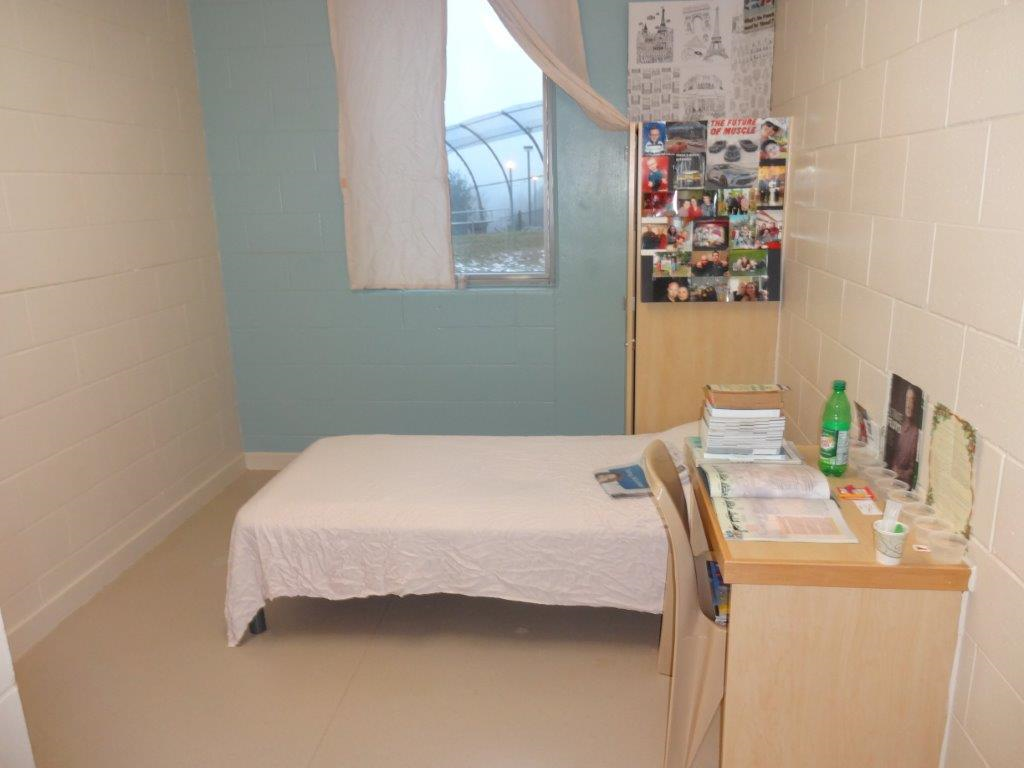 This image is of a standard single occupancy room at St. Lawrence Valley Correctional and Treatment Centre. It has an unobstructed window with curtains, a desk with moveable chair, and a personal wardrobe.