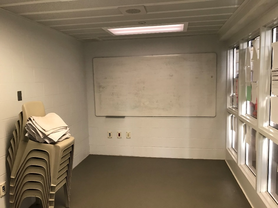 Cette image montre une salle de programme de groupe dans une unité de supervision directe avec un tableau blanc et des chaises en plastique empilées pour les détenus.