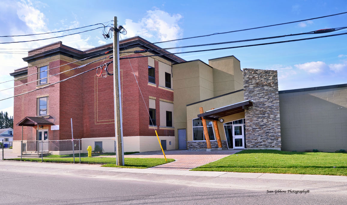 Photo of the Timmins Native Friendship Centre, located in a former school. It is a two-storey red brick building. Part of the building is stone. The entrance has a slanted roof held up by two wooden posts.